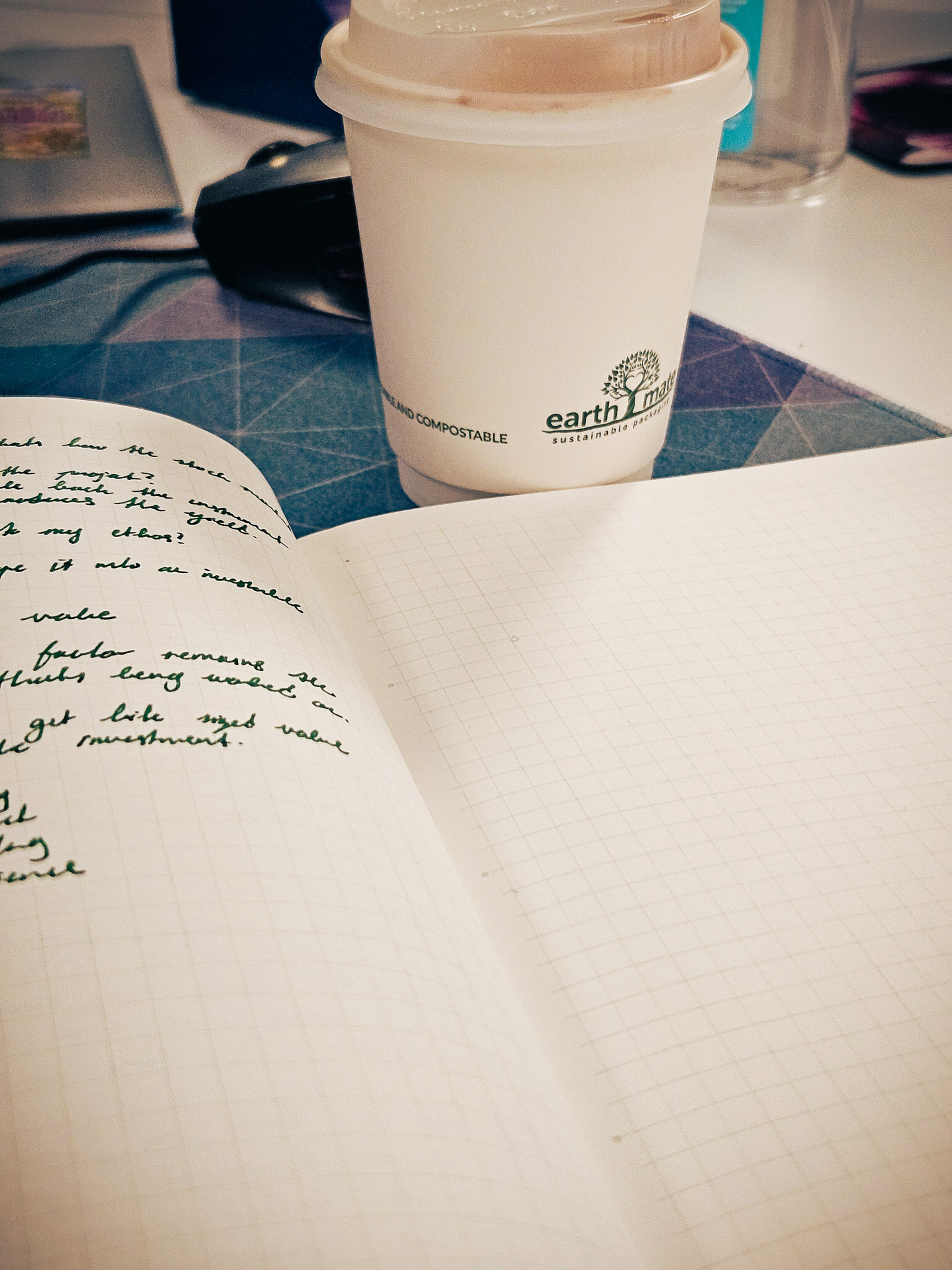 An open gridded notebook with handwritten notes beside a takeaway coffee cup on a desk.